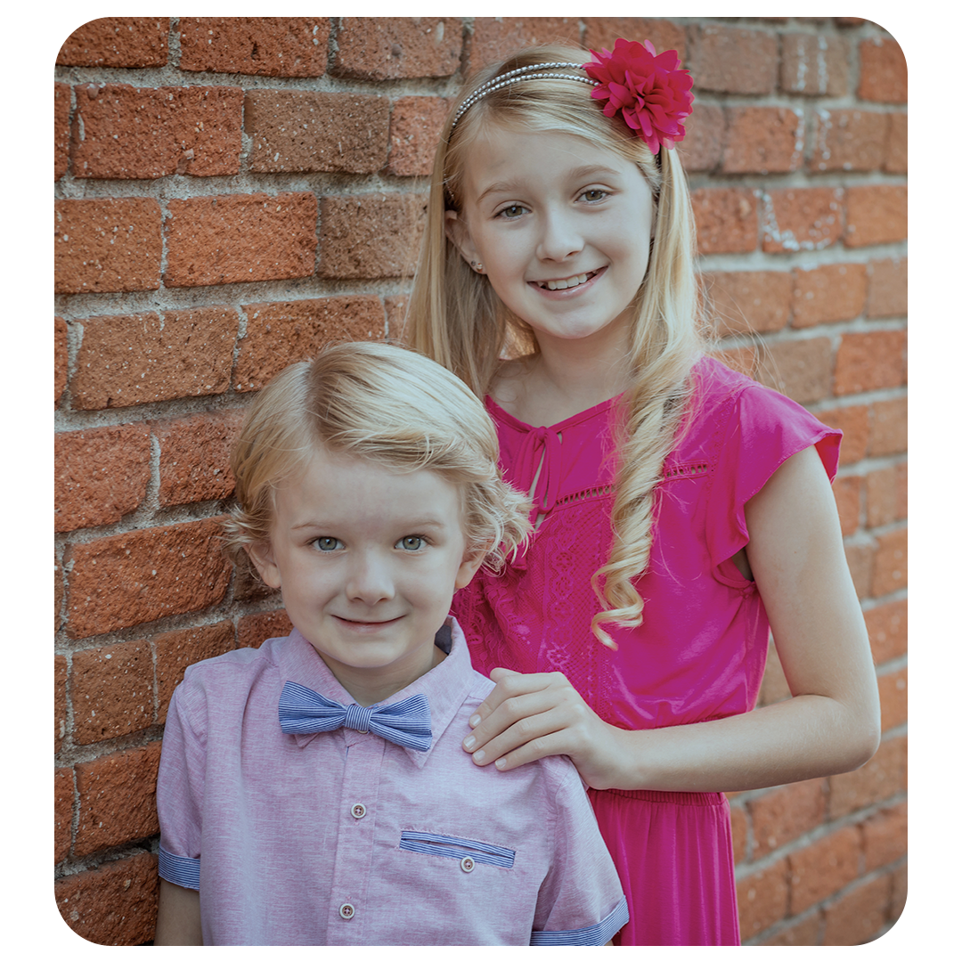 A natural light portrait of a brother and sister with blonde hair against a brick wall